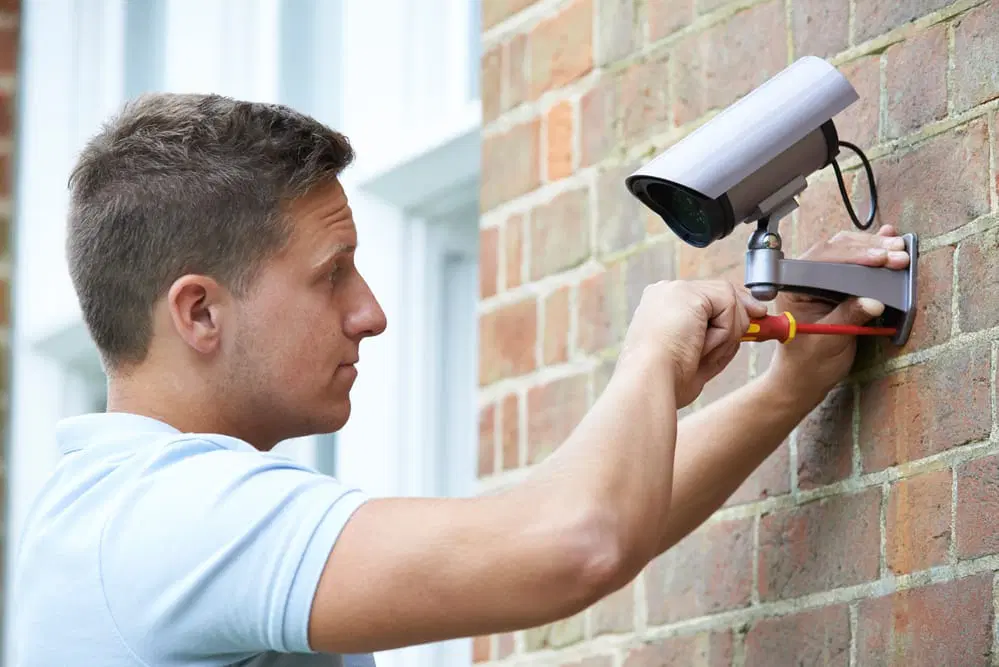 Man installing a security camera on a brick wall, using a screwdriver. The camera is mounted above a window, and he appears focused on the task.