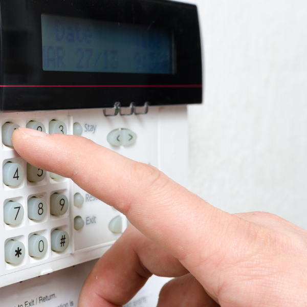 A person's hand pressing buttons on a security alarm keypad, displaying a digital screen with date and time information above the buttons.