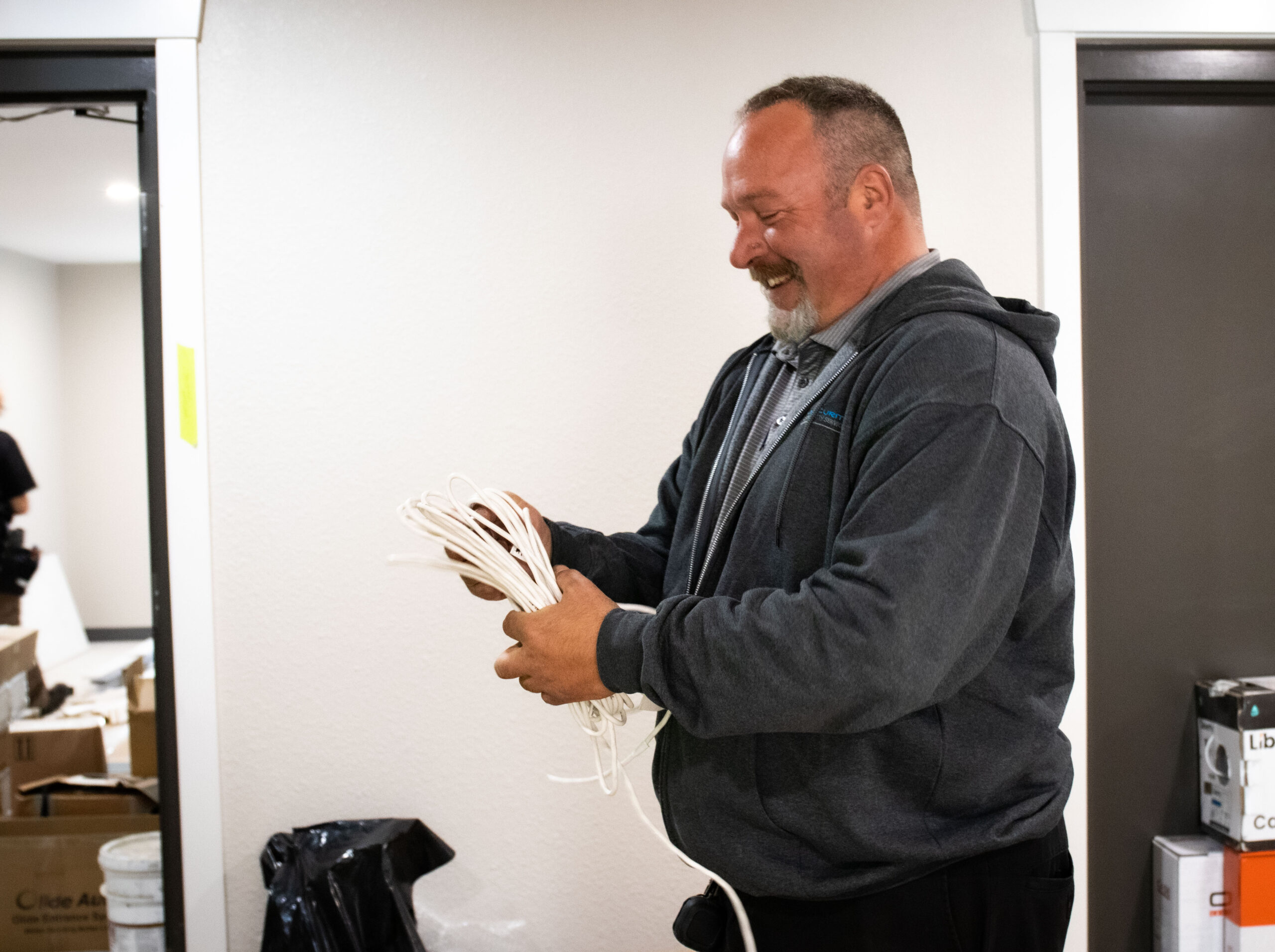 A man in a grey hoodie smiles while organizing white cables in a room filled with boxes and equipment.