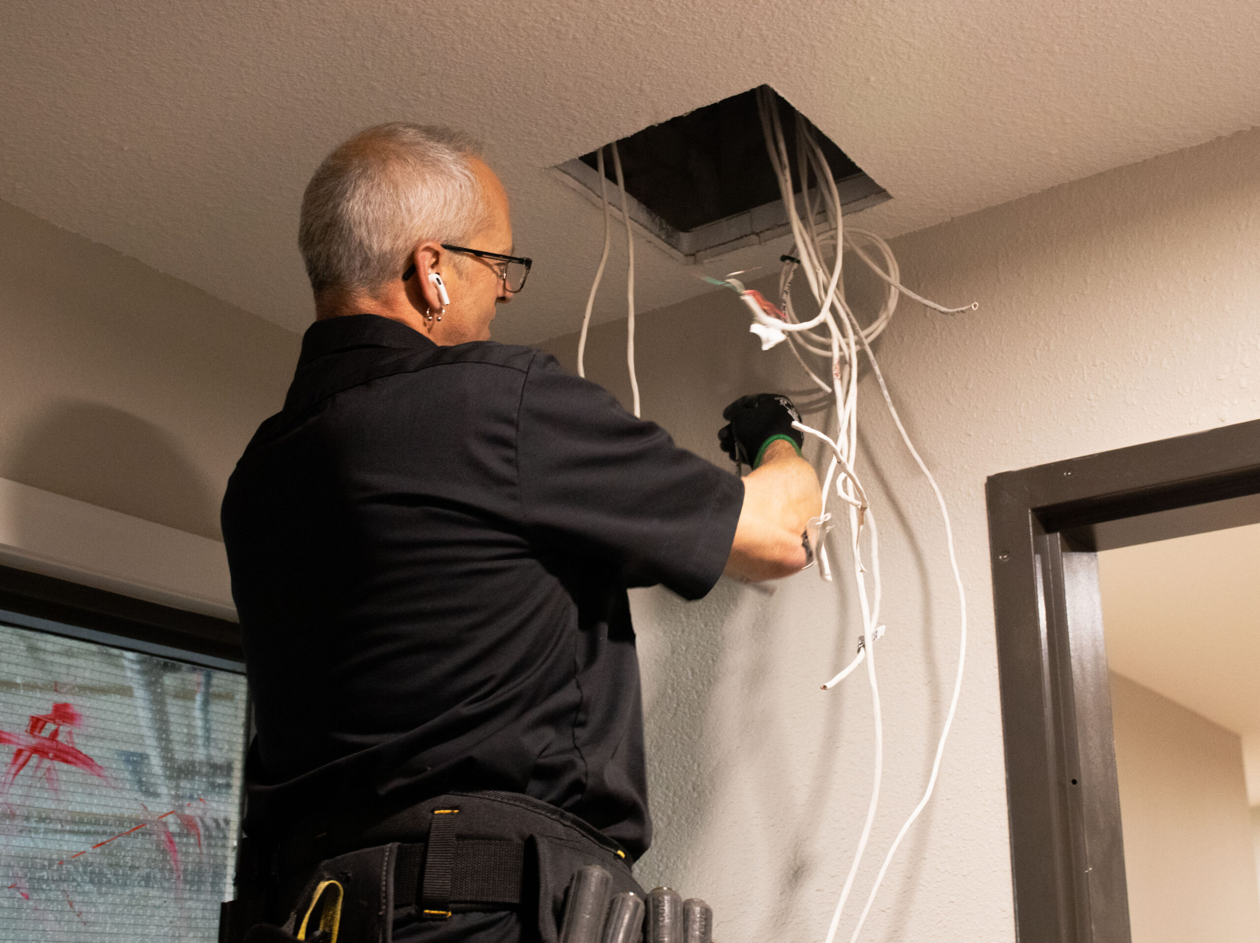 A person wearing a black shirt, earphones, and gloves is working on electrical wires coming from a ceiling opening, with tools in a belt.