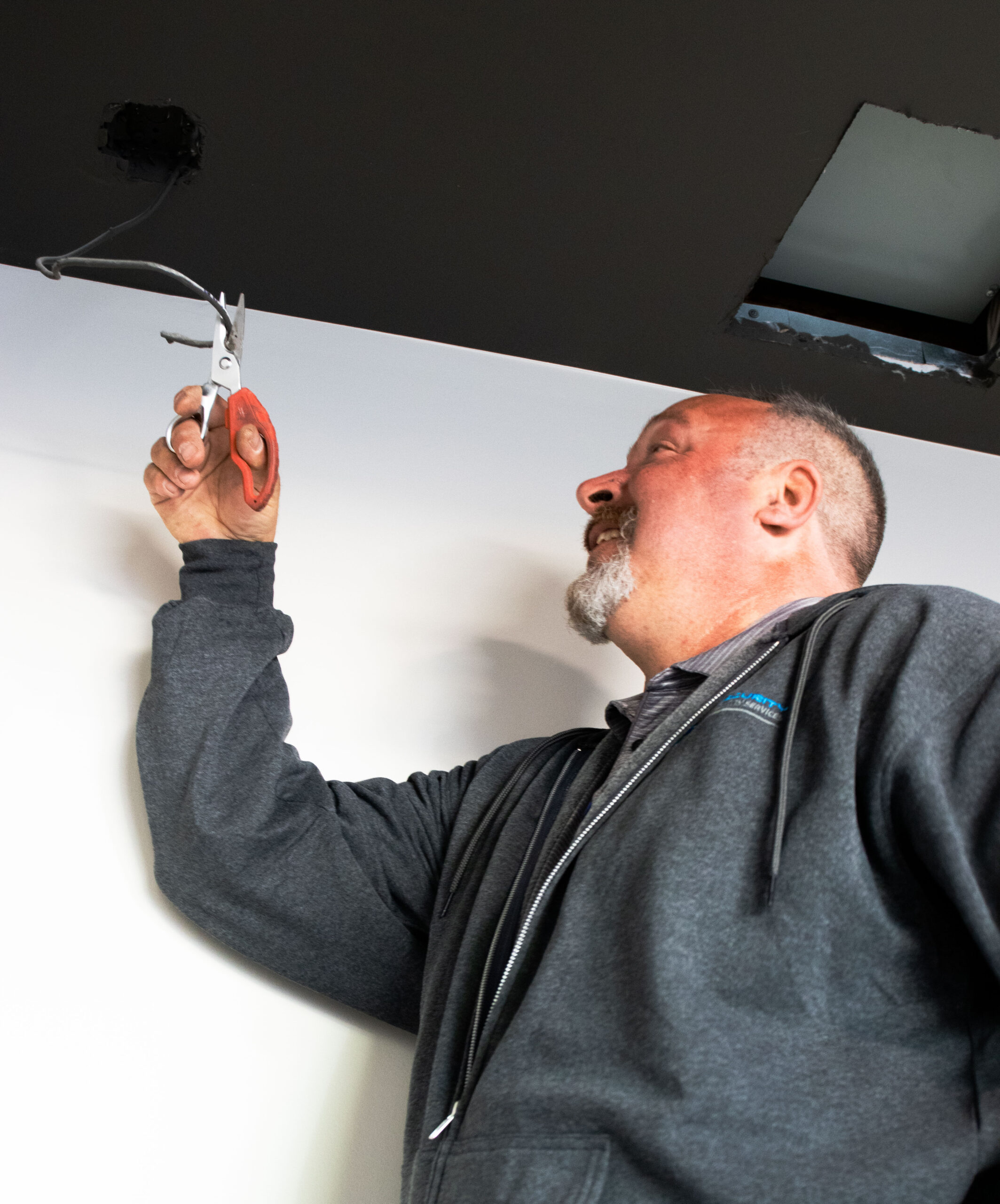 IMG_9450 A man in a dark hoodie trimming a wire hanging from a ceiling with scissors, looking upward intently.