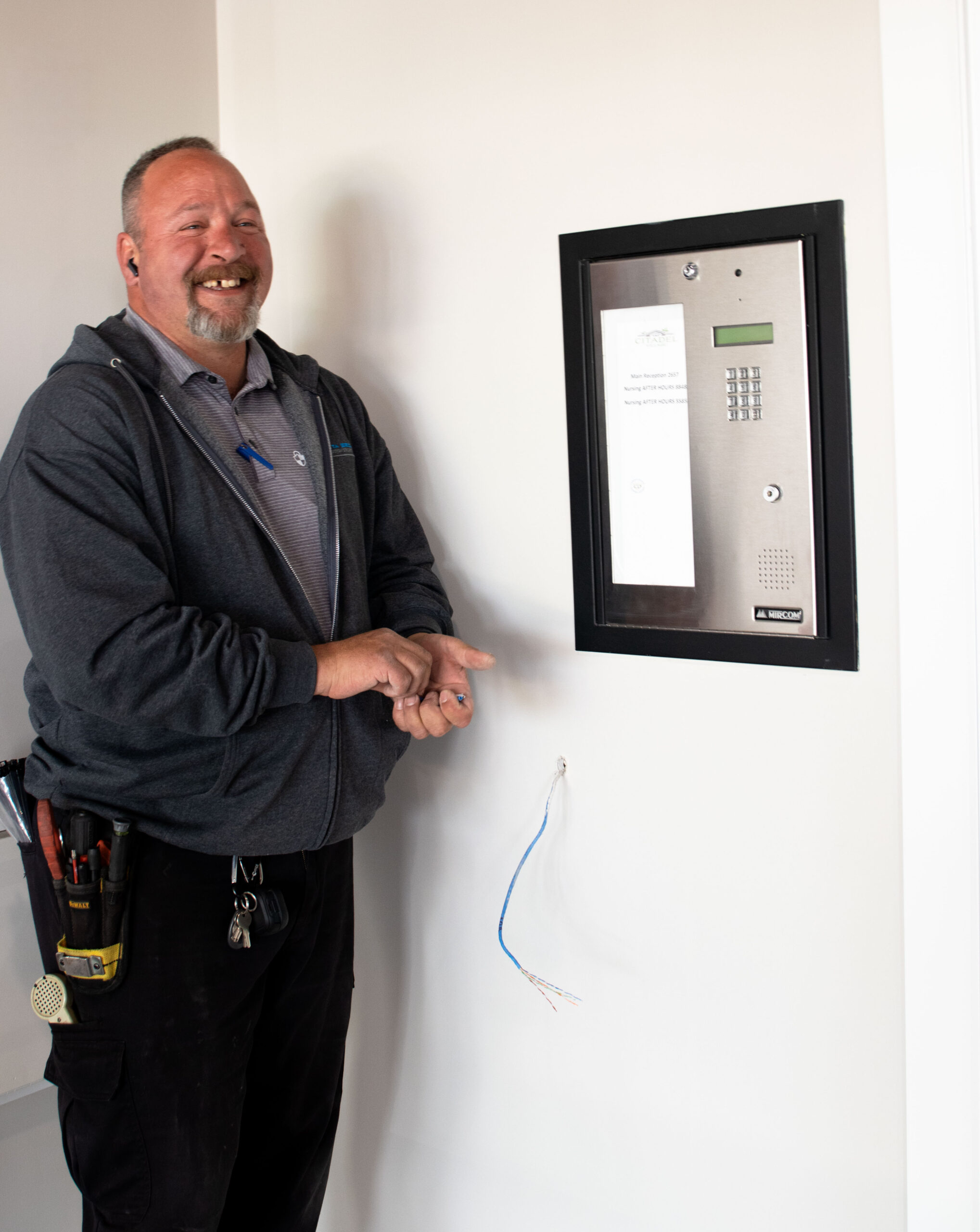 Smiling man in work attire stands next to an intercom system on a wall, holding wires. A toolbelt with work tools is visible at his waist.