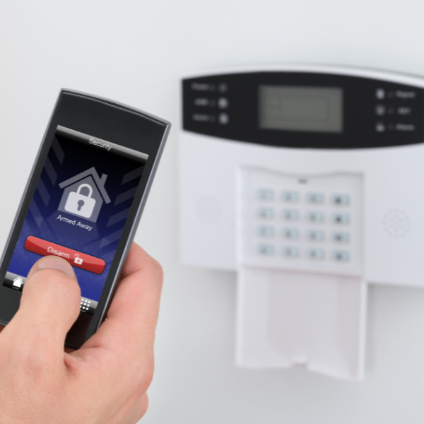 Close-up of a hand holding a smartphone displaying a home security app with a "Disarm" button. A blurred security keypad is in the background.