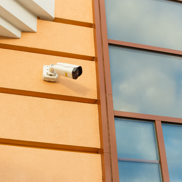 A white security camera mounted on an orange textured building wall, next to large reflective glass windows.
