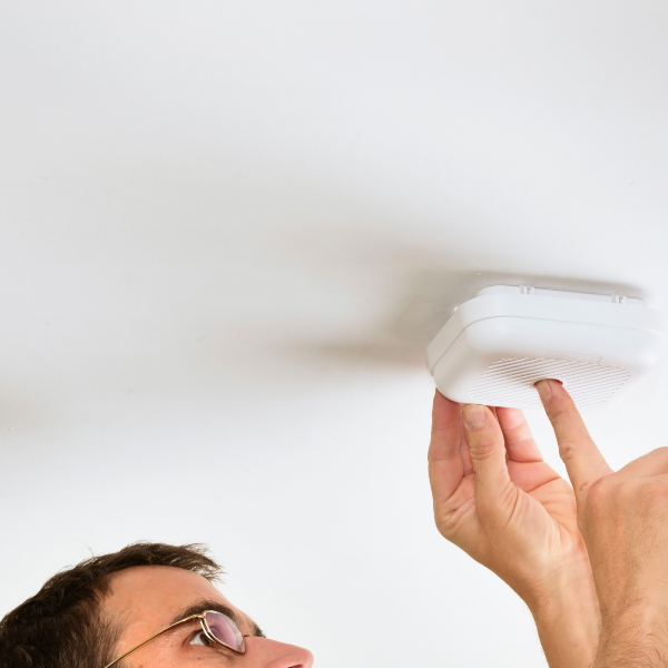 Person installing a white smoke detector on the ceiling, using their left hand to secure it in place. Only their forehead and hand are visible.